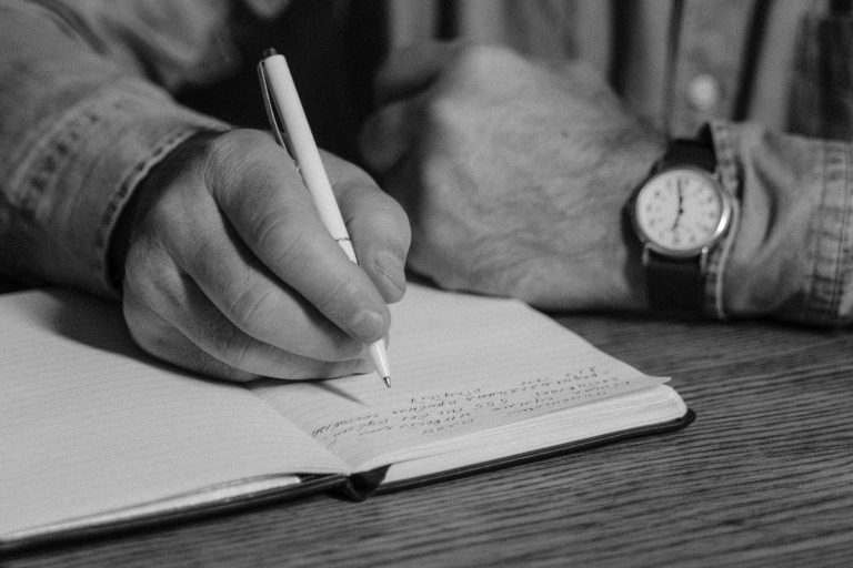 Black-and-white close-up of person writing in a notebook, emphasizing handwriting and detail.
